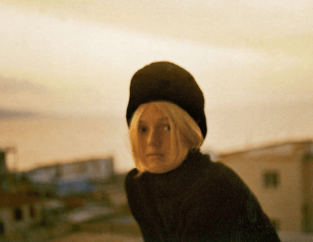 Young blond woman in a black fur hat and black turtleneck sweater with hazy houses and ocean in sunset background.