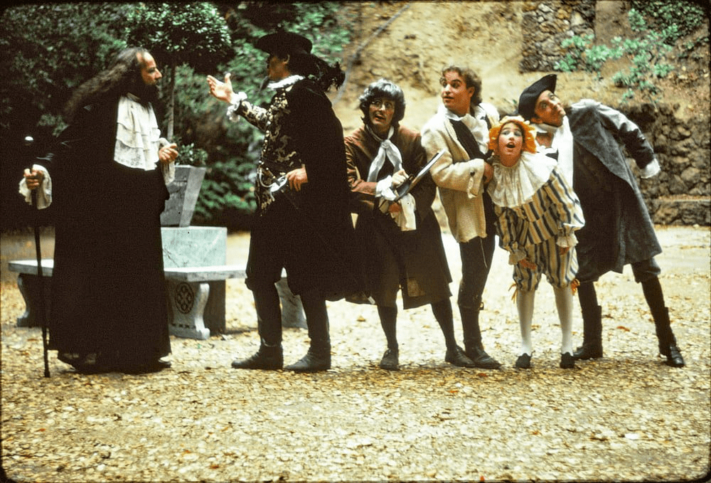 A group of five actors in Elizabethan costume lined up and listening in a Shakespearean comedy let outdoors with autumn leaves on the ground.