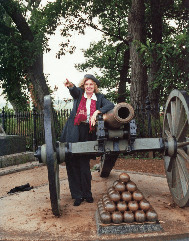 woman in a long gray coat and gray beret standing by an old Civil War cannon with a stack of cannon balls. She is pointing to something in the distance.