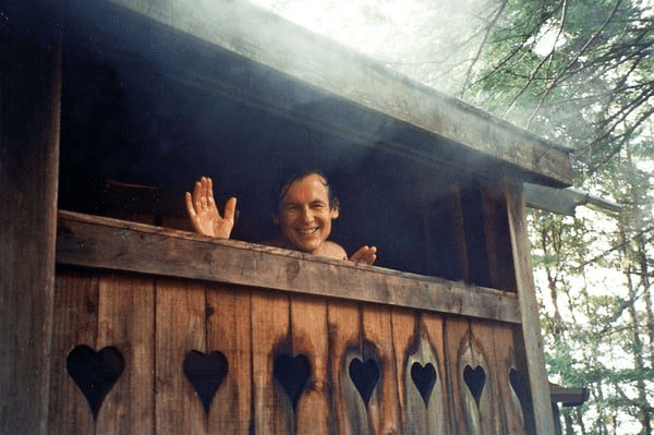 Smiling young man waving from inside a rustic wooden outdoor shower in the forest.