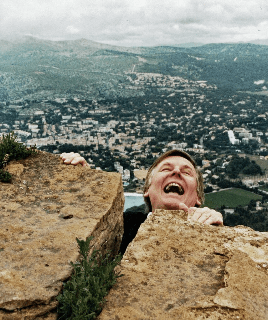 Man pretending to scream while hanging by his fingers to rocks over a landscape of green hills and houses. Meant to be a funny image.