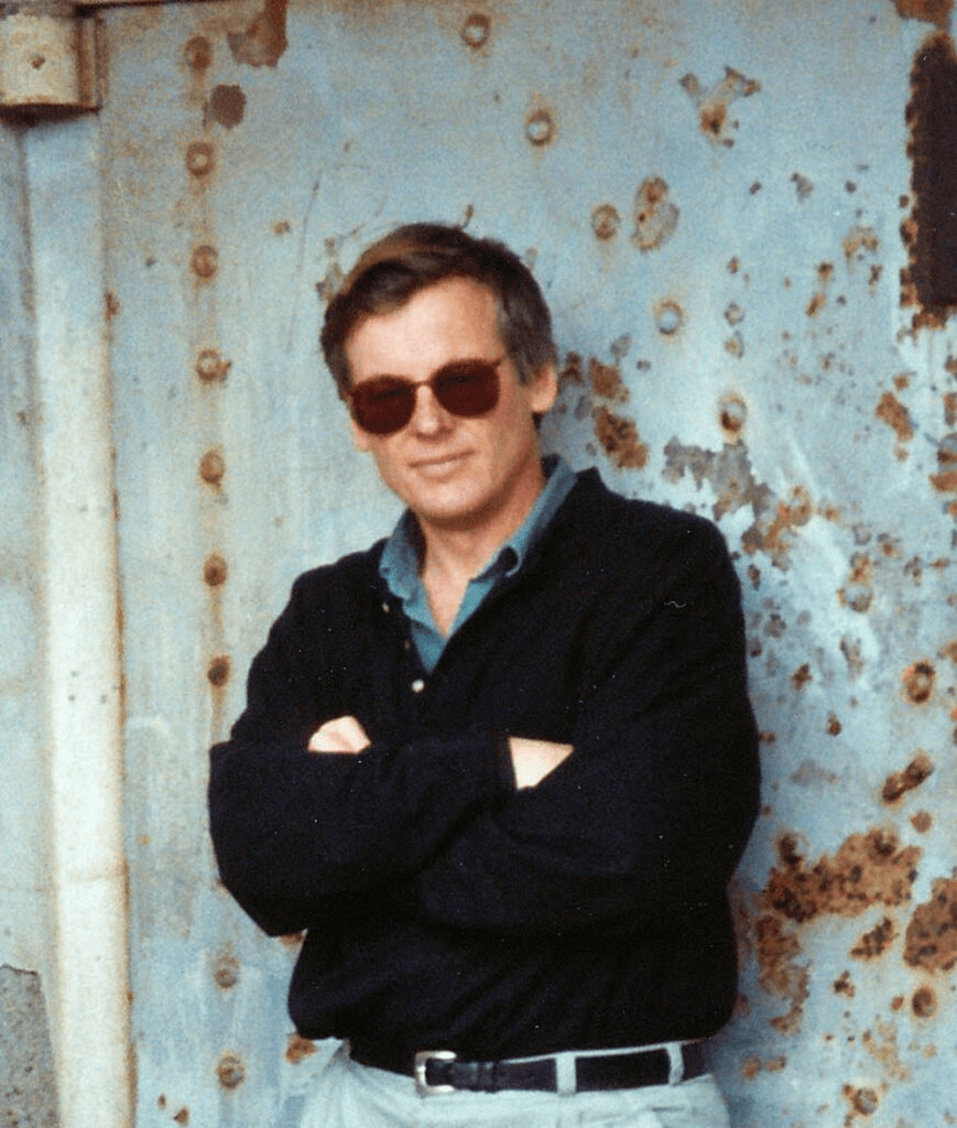 Young man wearing dark glasses and a black shirt against a pale blue wall with rusty accents.