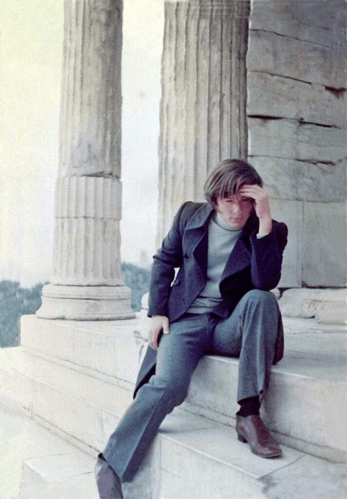 Young man in long coat, sitting on steps of a Greek temple and thinking with his hand to his head.