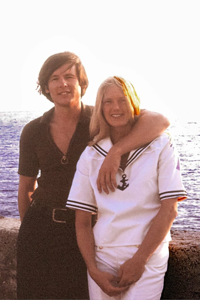 Attractive young couple. Man wearing brown v-neck shirt with his arm around blond woman in a sailor suit. They are standing by an old stone wall with the ocean in the background.