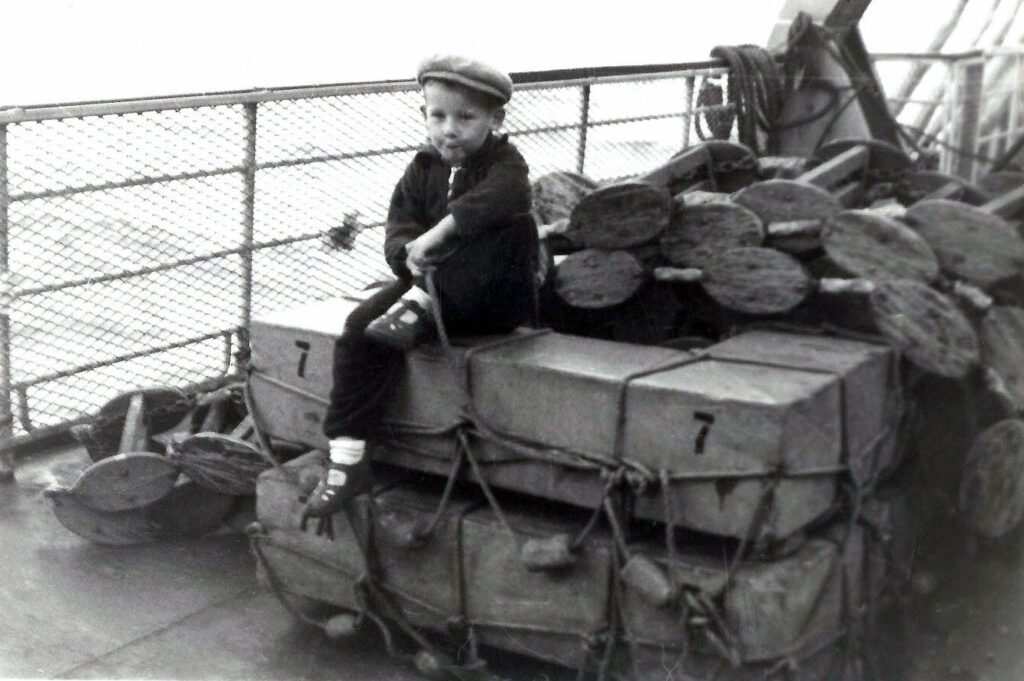 Small refugee boy sitting on boxes onboard a ship heading to America in 1949.