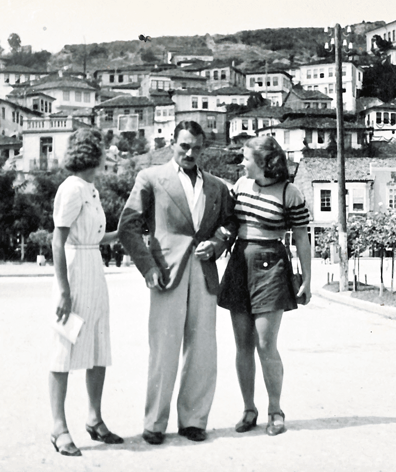 Black and white photo of two women holding the arms of handsome man with moustache wearing a suit with a European hillside village in background.