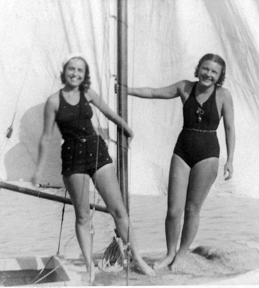 Black and white photo of two young women wearing bathing suits and holding onto the mast of a sailboat.