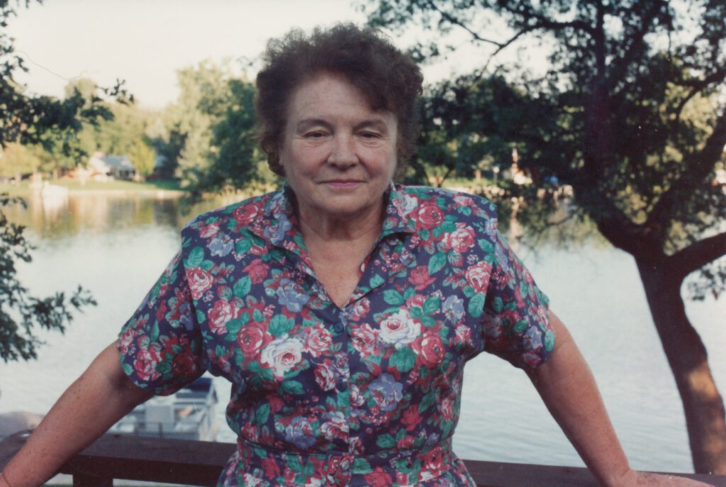 Older woman in flowered dress with trees and lake in background.