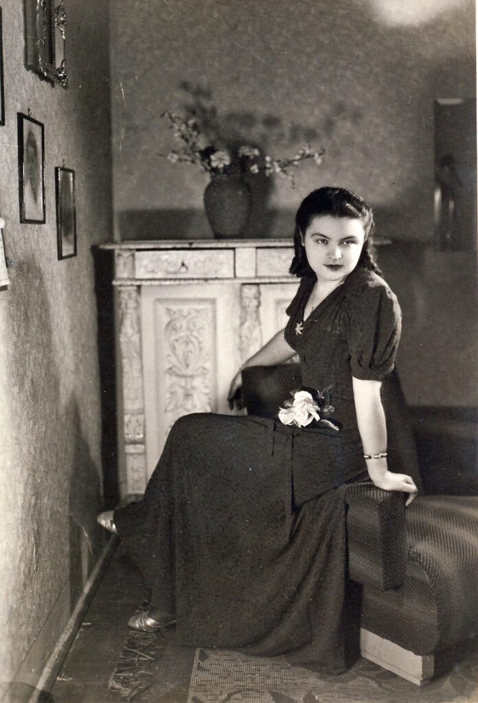 Glamorous young woman in 1940 wearing an evening dress and sitting on a sofa arm in an old fashioned room.