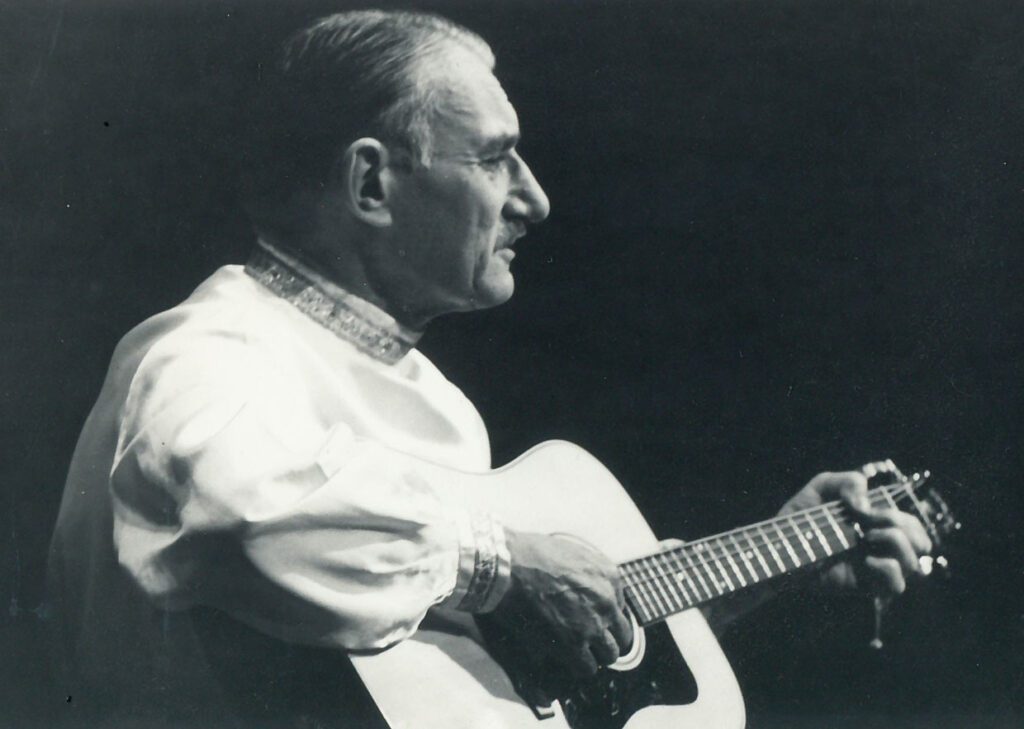 Older man wearing a Russian shirt and playiing a guitar.