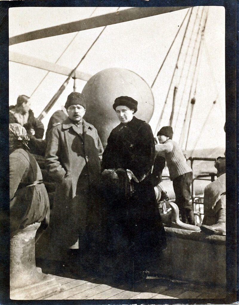 Older refugee couple in winter coats and hats onboard a ship with other refugees in background.