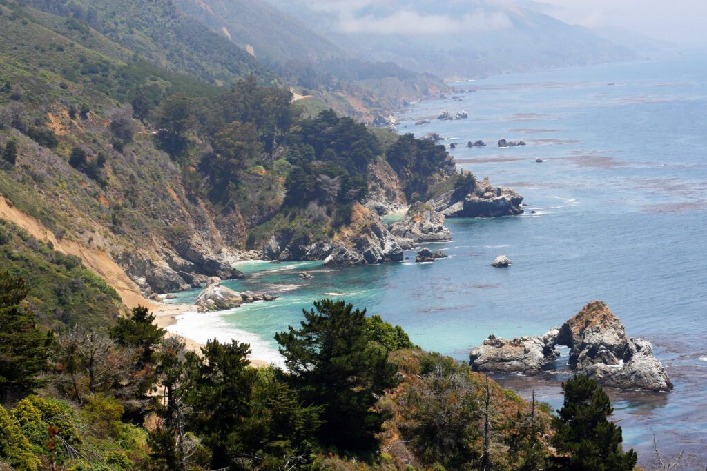 California coast view of steep cliffs, pine trees, and rocky ocean shore.