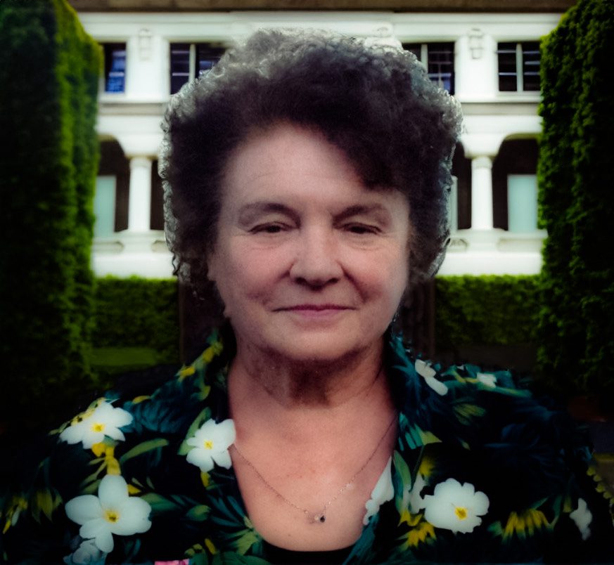 Smiling older woman wearing a flowered shirt with trees and college campus in background.