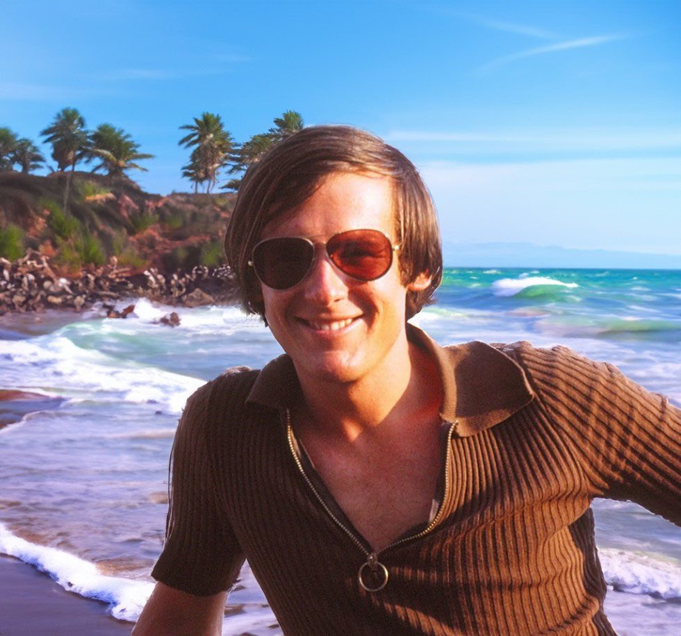 Young man by the seaside wearing a brown shirt and sunglasses. He's grinning.